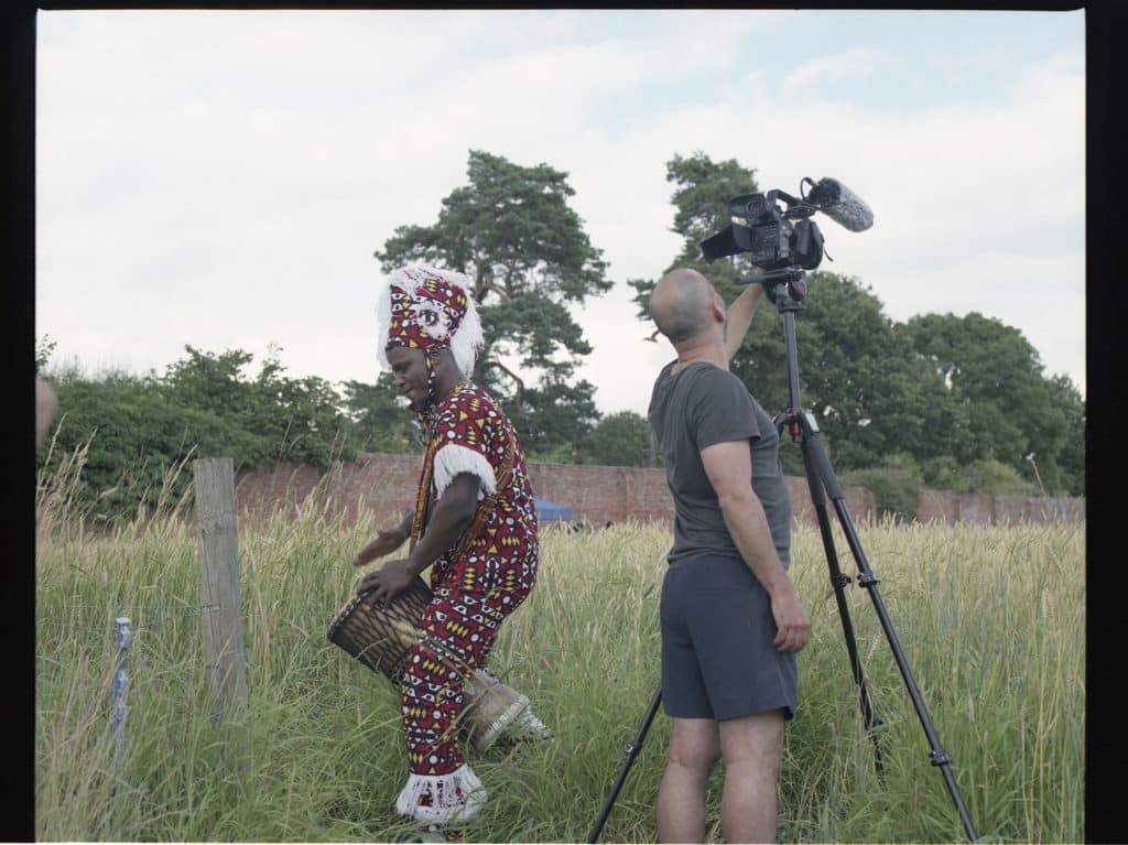 African drummer Abraham Paddy Tetteh with camerman Rui Pignatelli in a field of wheat during filming for Hand, Earth, Gesture, Return (wheat field drumming jam), Hatton Farm, Warwickshire, 2021