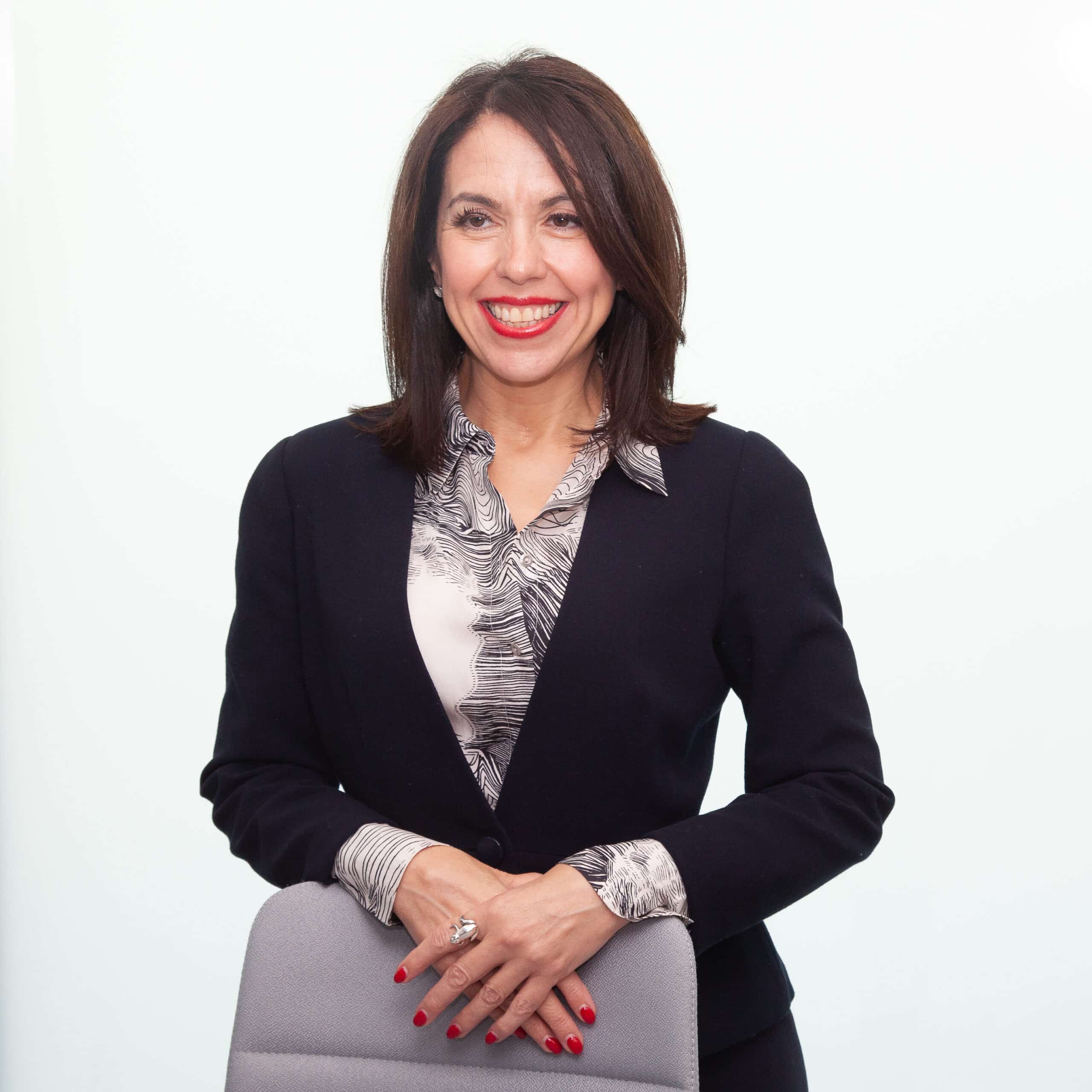 Portrait of Hispanic lady with shoulder-length brown hair, standing behind a chair.